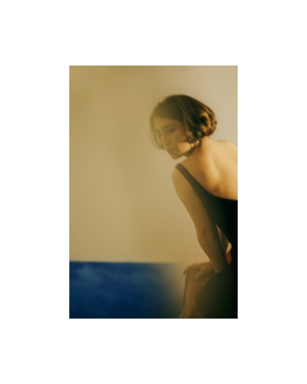 A dark-haired figure indoors, partly obscured by blurred shapes and patches of colour.
