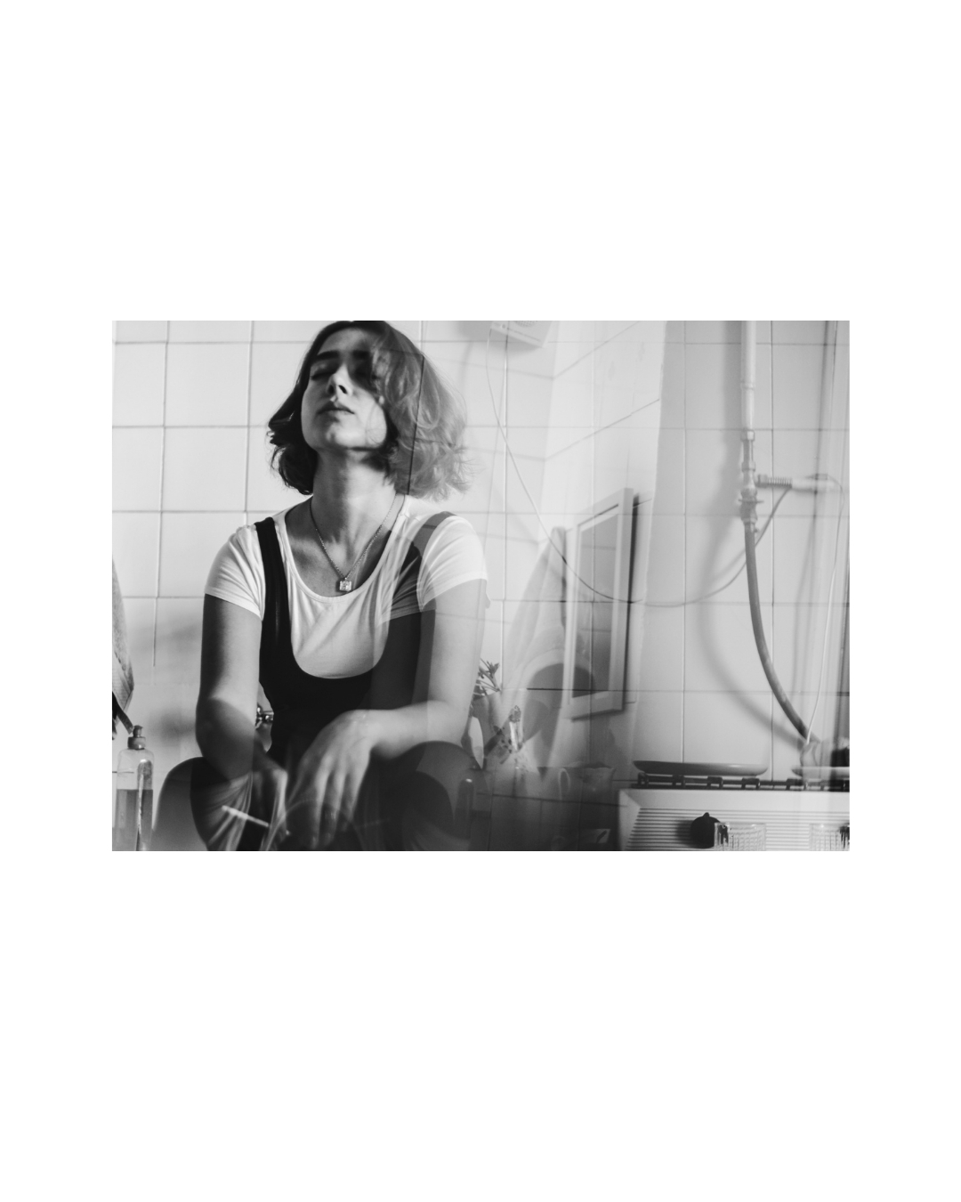 Black-and-white view of a woman seated against tiled walls, with dark straps over a white T-shirt.