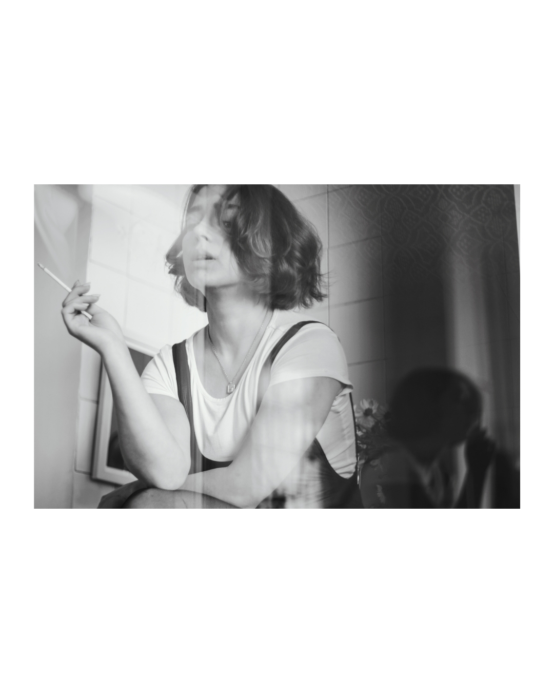 A seated woman with short dark hair and a white T-shirt in a tiled kitchen, shown in black and white.