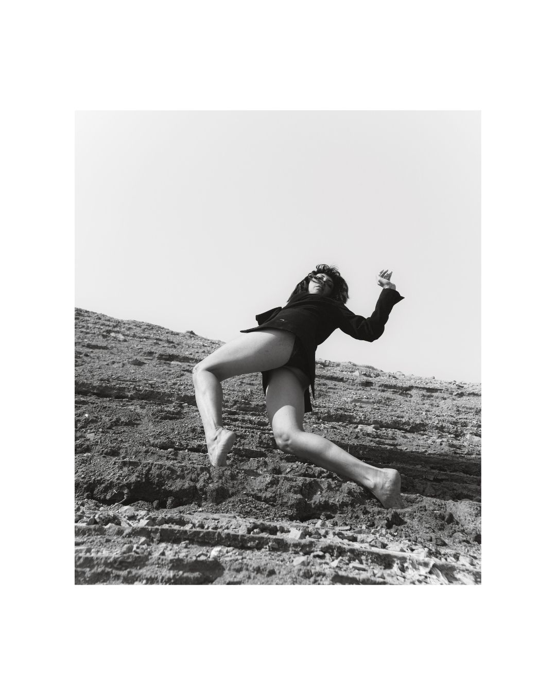 A woman near water in an open, rocky setting, with daylight and shadow defining her figure.
