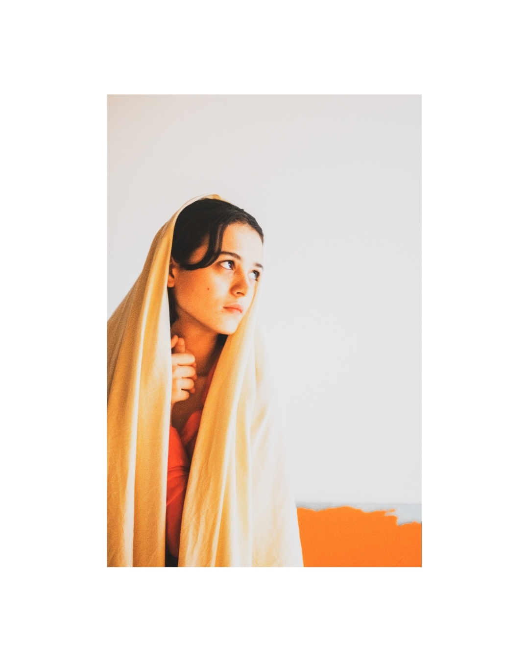 A woman in a red-orange dress poses against a pale wall in a softly blurred colour photograph.