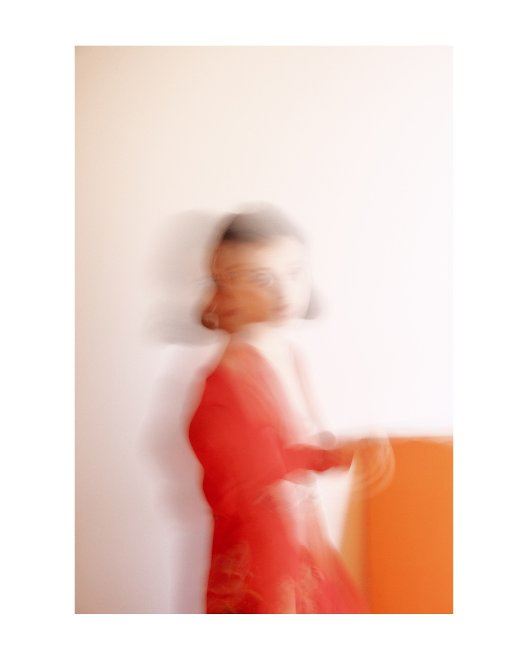 A woman in a red-orange dress poses against a pale wall in a softly blurred colour photograph.