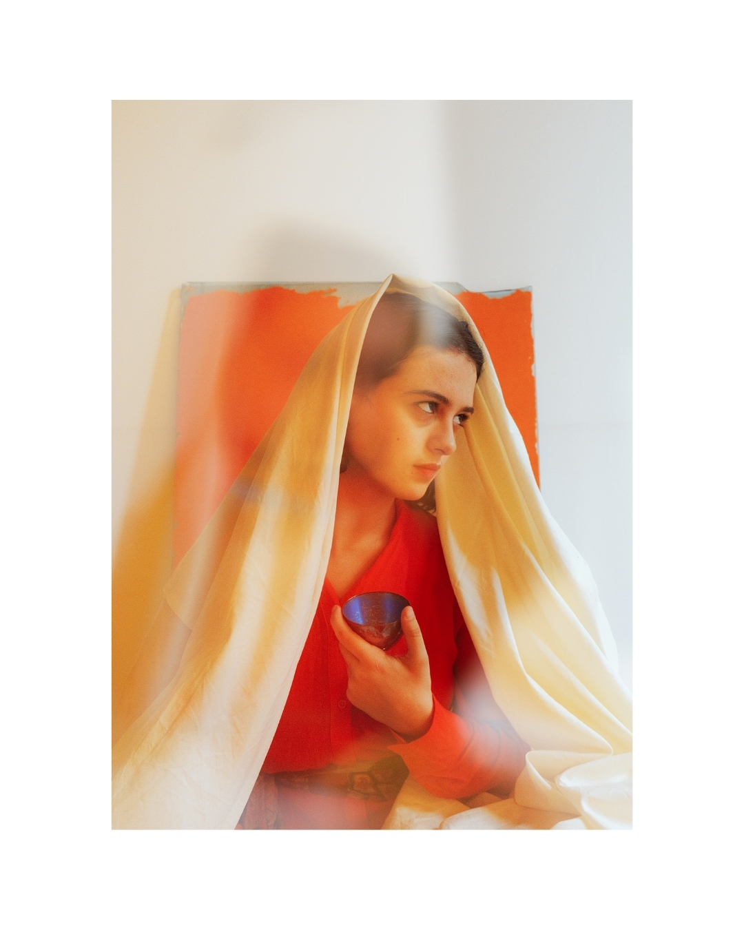 A woman in red-orange clothing appears in soft focus in a bright room with pale walls.