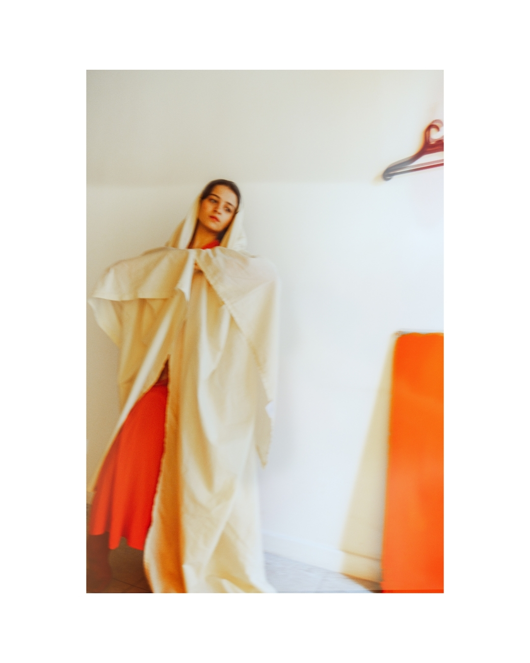 A softly blurred woman in red-orange clothing against a pale wall, by Lilit L Hovhanni.