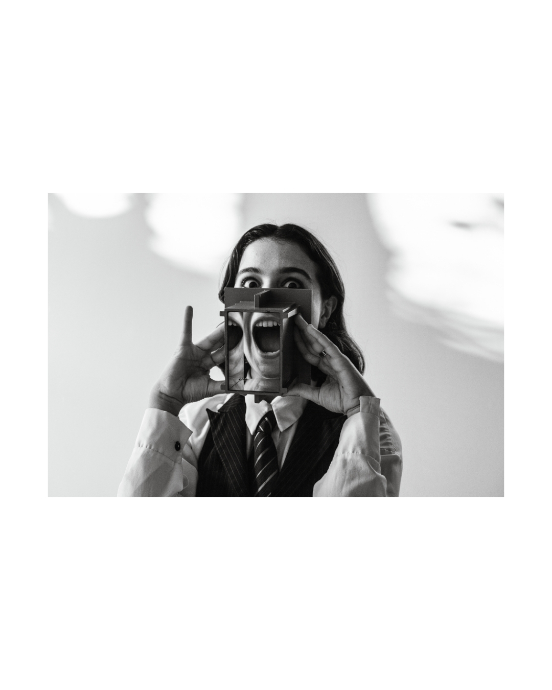 Hands hold a rectangular frame around parts of a face against a light backdrop in grayscale.