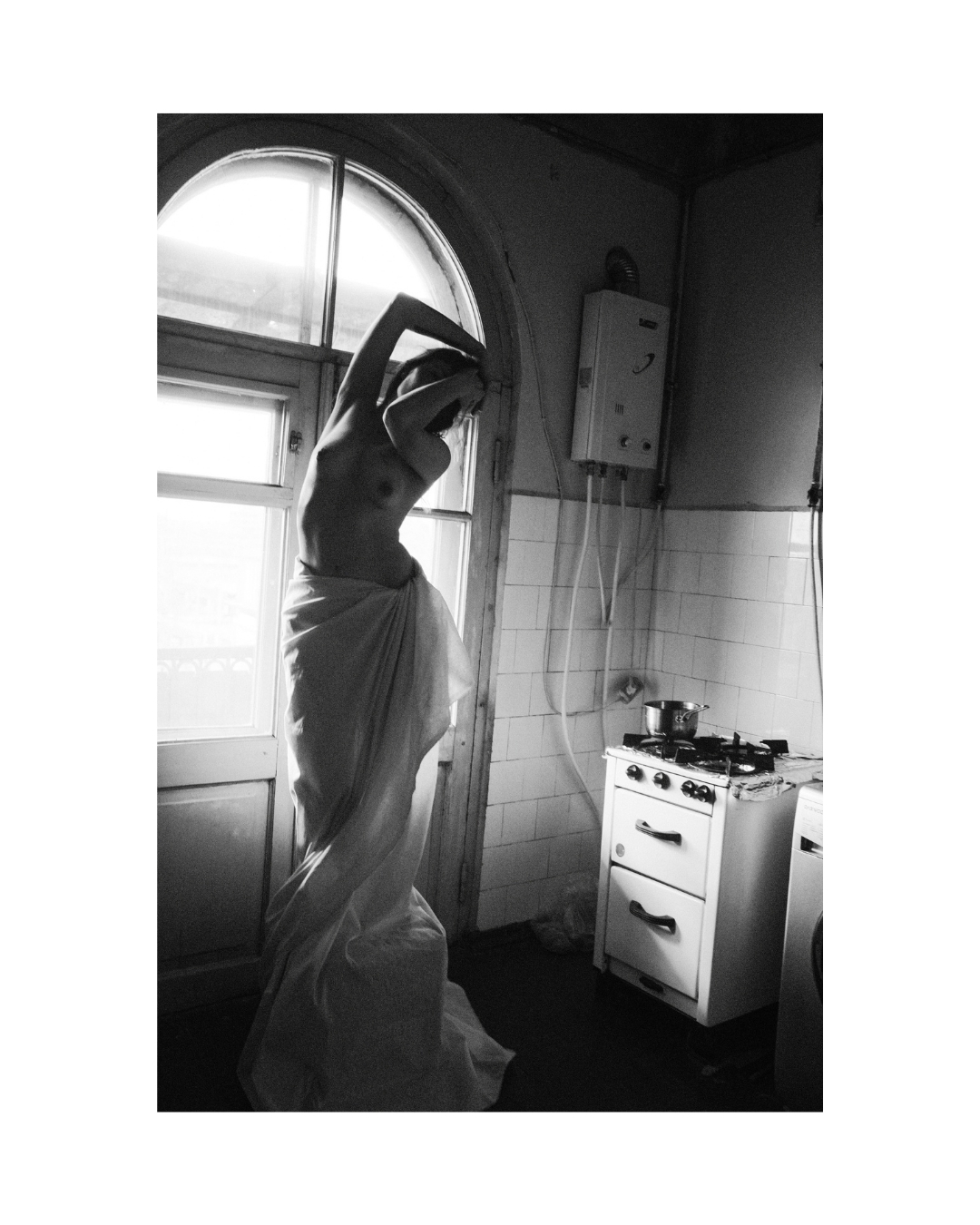 Black and white view of a bare-chested figure in light fabric, posed in a dim interior with bright window light.