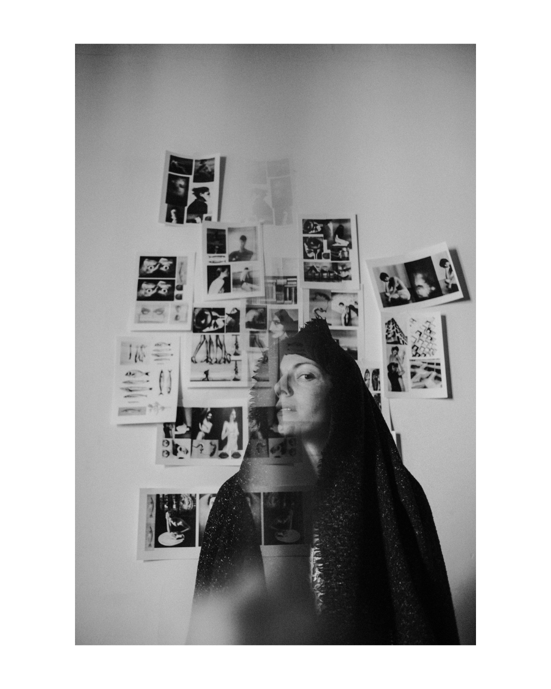 A figure poses indoors before a wall of printed pictures, in black and white.