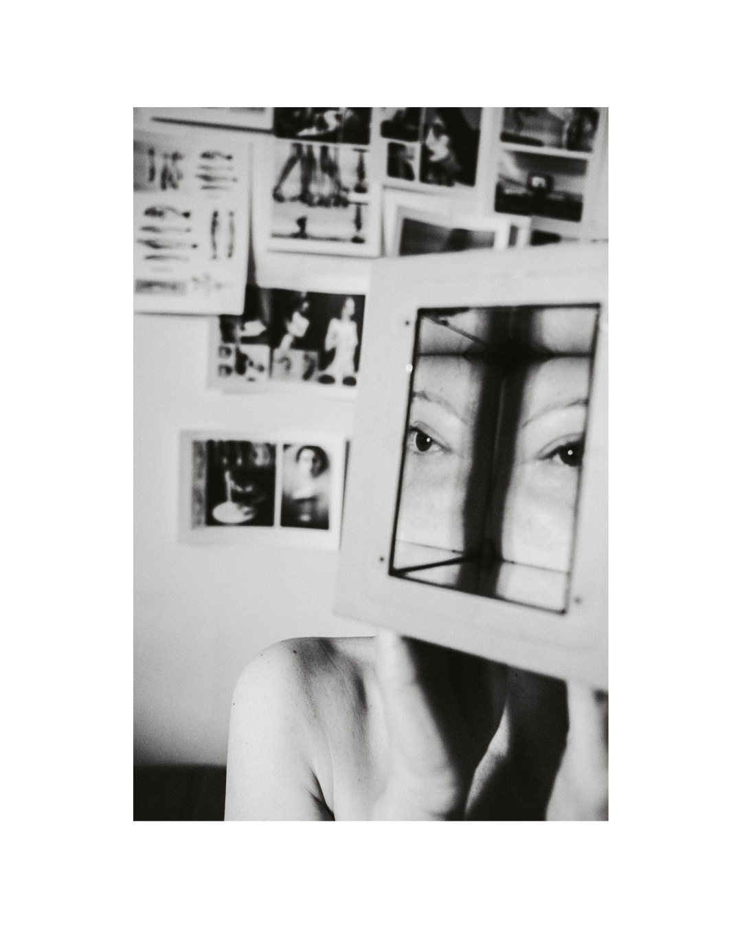 Black-and-white portrait of a figure with small printed images arranged on the wall behind them.