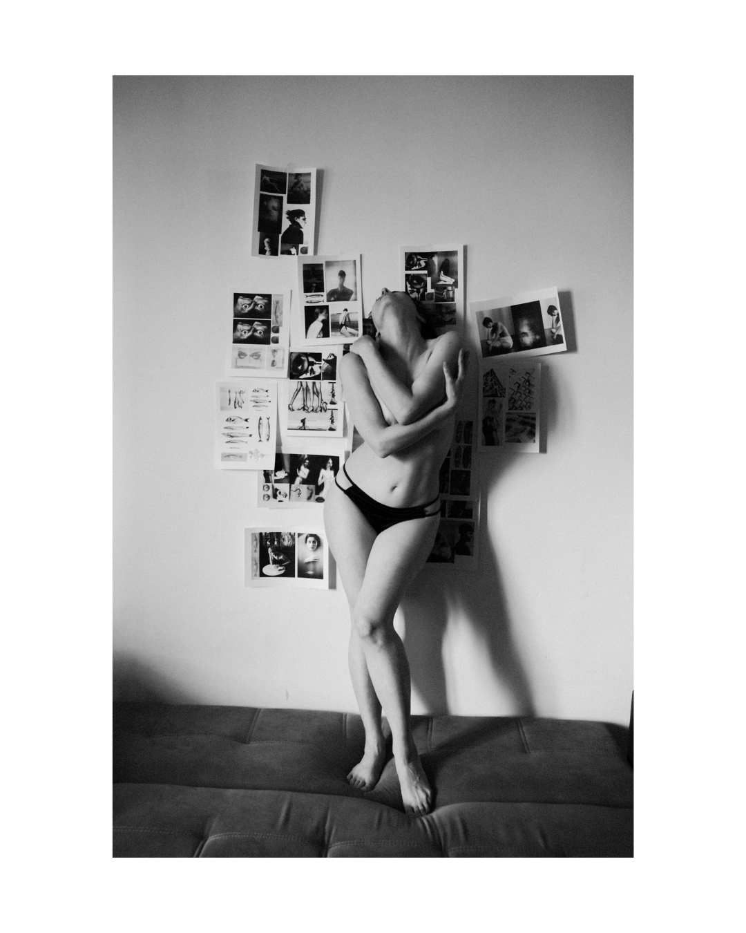 A figure in light and shadow against an indoor wall covered with printed pictures, in black and white.