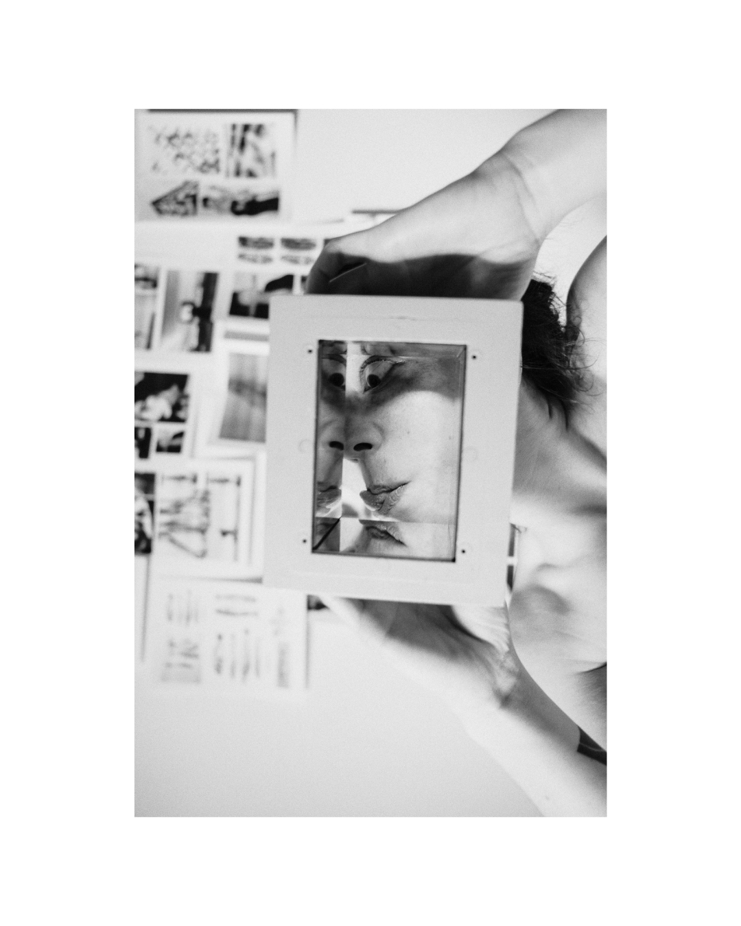A figure poses indoors before a wall of printed pictures, in black and white.