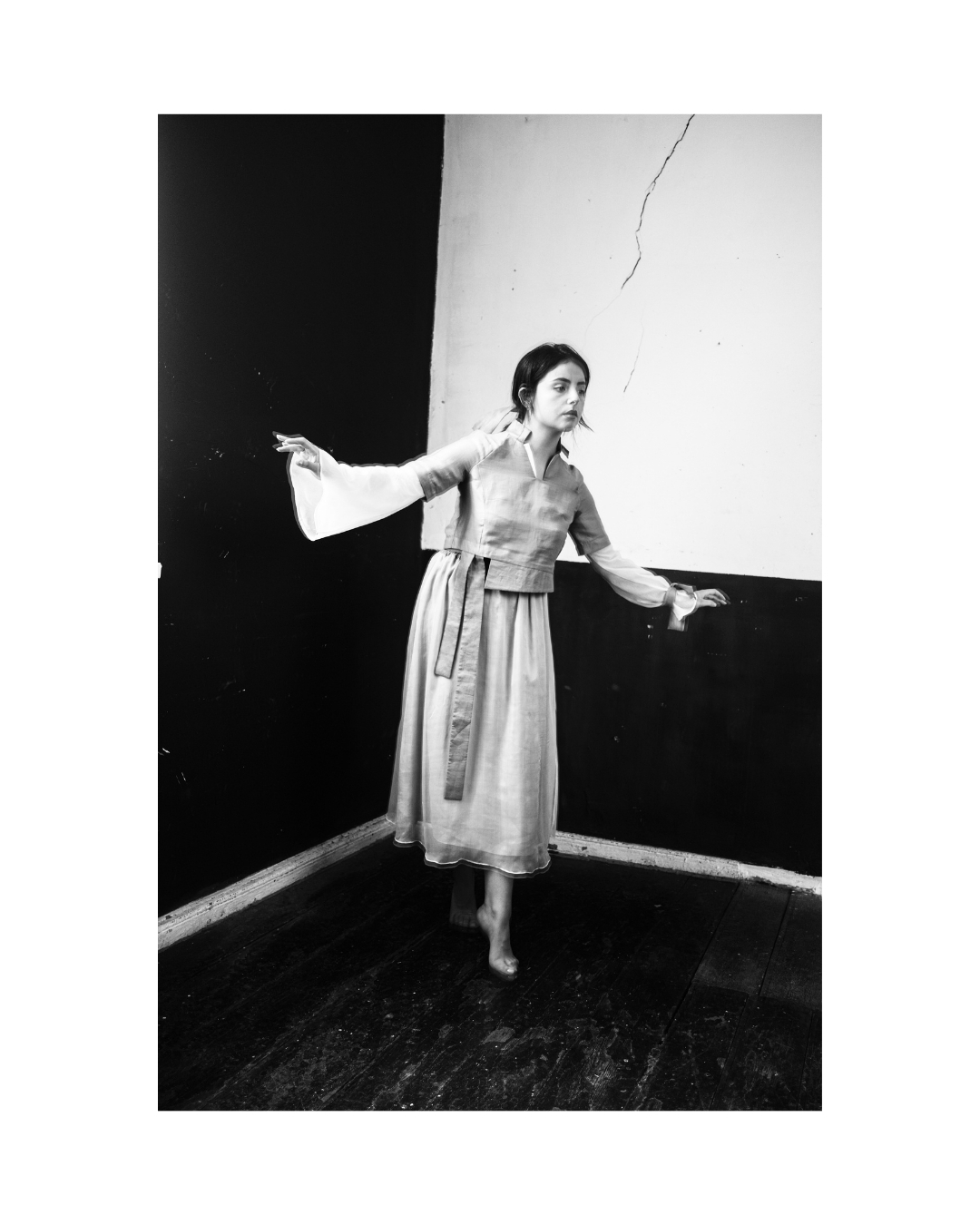 A figure in a light dress contrasts with a dark interior wall in black and white.