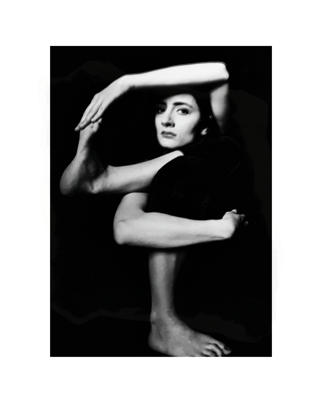 A figure with folded limbs and dark clothing against a black background, photographed in black and white.